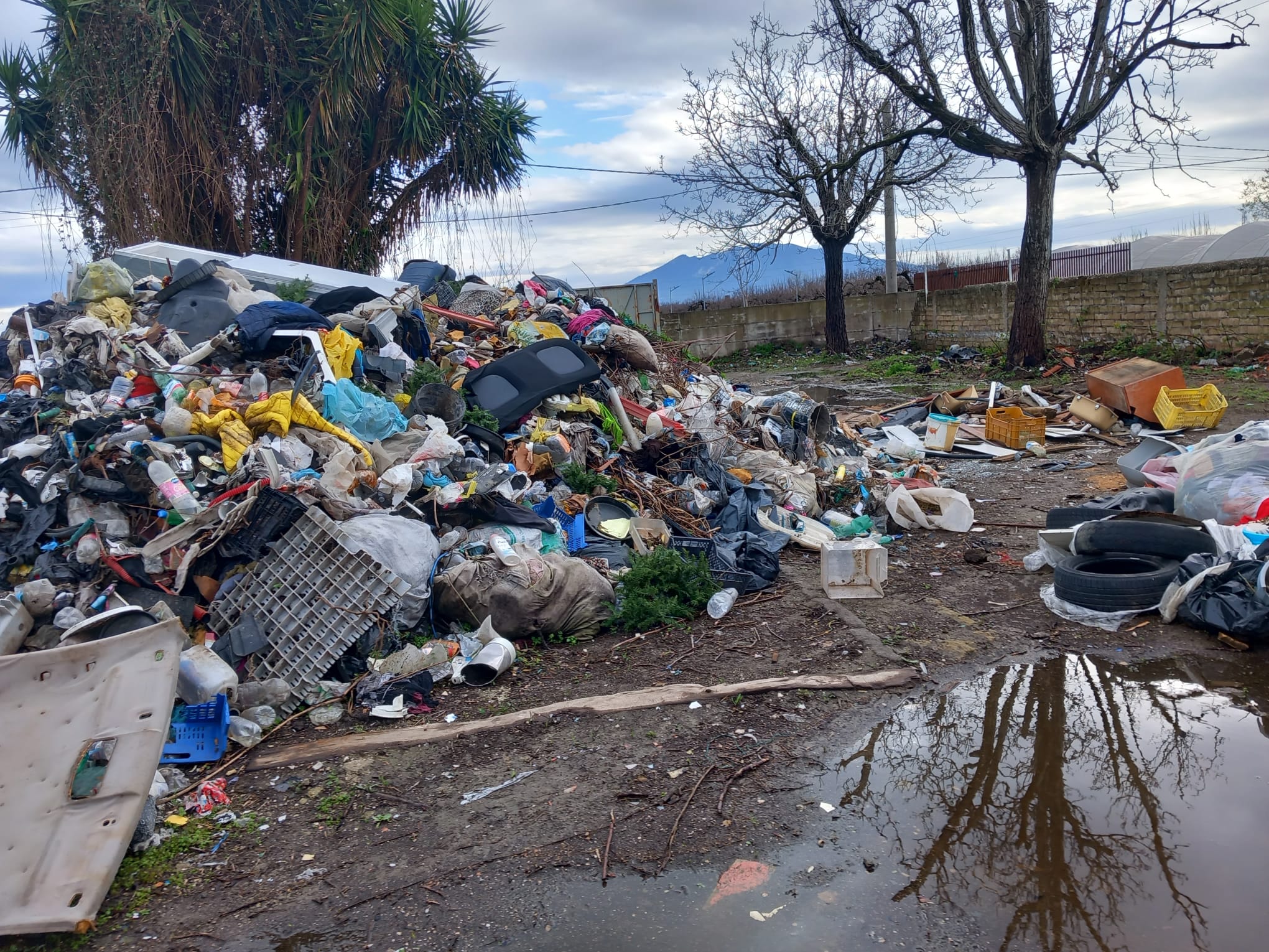 Discarica - Un grande mucchio di rifiuti assortiti, plastica e detriti è sparso su un terreno fangoso vicino ad alberi spogli, con una pozzanghera che riflette la scena. Ci sono cieli nuvolosi e una recinzione sullo sfondo.
